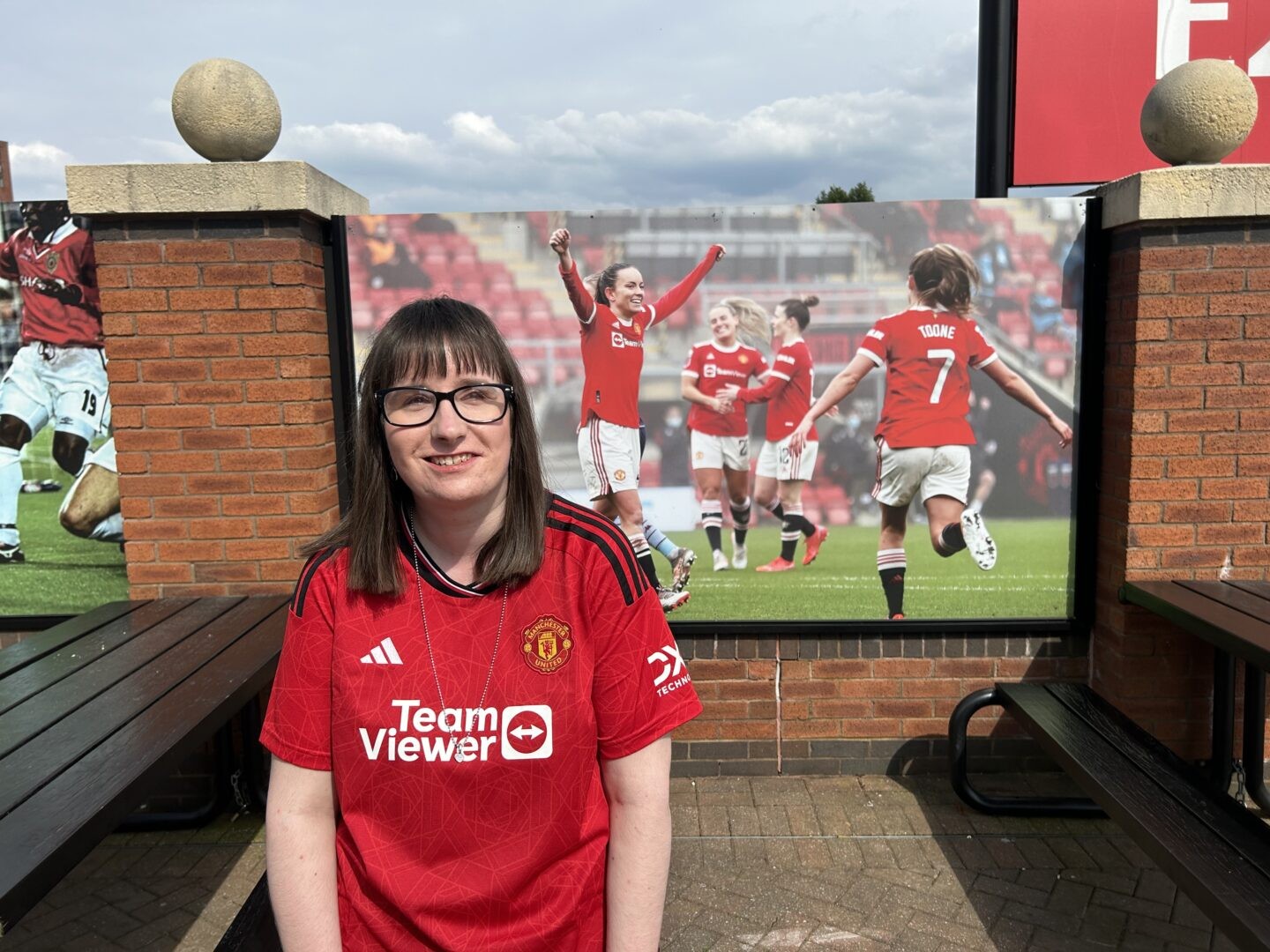 Holly, mặc áo Manchester United màu đỏ, đang đứng trước một tấm áp phích có ảnh 4 cầu thủ Manchester United Women đang ăn mừng một bàn thắng.