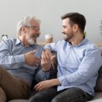 Người đàn ông lớn tuổi đang ngồi trên диван nói chuyện với con trai đã trưởng thành của mình.
